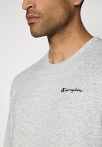 Light grey crewneck t-shirt made of soft fabric, featuring a blue embroidered "Champion" logo on the chest and a ribbed neckline.