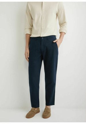 Mężczyzna ubrany w beżową koszulę z długim rękawem, ciemnoniebieskie spodnie na miarę oraz brązowe zamszowe mokasyny z jedną ręką w kieszeni na jednolitym tle.