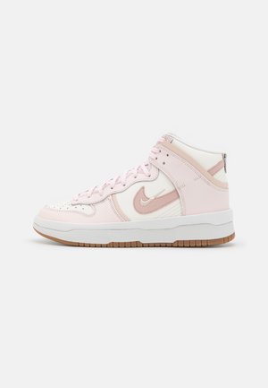 Valkoisen ja vaaleanpunaisen korkeavartinen lenkkari, jossa Nike-logo, edessä nauhat ja ruskea kumipohja, yksinkertaisella vaalealla taustalla.