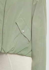 Giacca bomber verde chiaro con chiusura a zip, orlo elastico e una tasca laterale con bottone a pressione. Texture liscia, design minimalista.