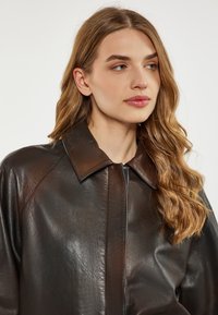 Braune Lederjacke mit großem Kragen und Reißverschluss. Das strukturierte Material hat einen leichten Glanz, der die satte Farbe der Jacke unterstreicht.
