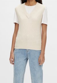 Personne portant un gilet en maille sans manches crème par-dessus une chemise blanche à manches courtes avec un jean en denim bleu clair, visible du cou aux hanches.