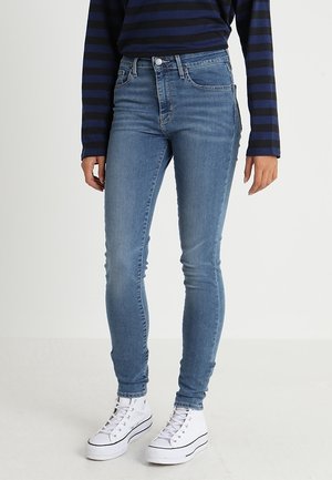 Person trägt hellblaue enge Jeans, weiße High-Top-Sneaker und ein langärmliges T-Shirt mit navyblauen und schwarzen Streifen.