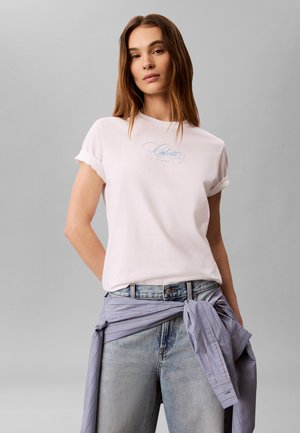 Junge Frau mit langen braunen Haaren, die ein weißes T-Shirt mit hochgekrempelten Ärmeln und hellblaue Jeans trägt, mit einem gestreiften Hemd um die Taille gebunden.