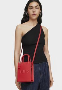 Bolso de mano de cuero rojo con logo "TOUS" en relieve, forma rectangular, asas cortas y correa de hombro desmontable. El modelo lleva una blusa negra.