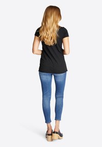 Kvinde med langt blondt hår iført en sort T-shirt, blå skinny jeans og sorte træsko med træsål, stående med ryggen mod.