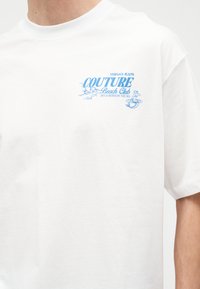 Λευκό μπλουζάκι με μπλε κείμενο "Versace Jeans Couture Beach Club" και κύματα, που εμφανίζει μια μικρή εικονογράφηση γοργόνας στο στήθος.