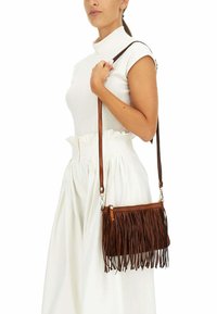Borsa a tracolla in pelle marrone con dettagli in frange, chiusura con zip e tracolla regolabile. Indossata sopra una maglietta e una gonna bianche.