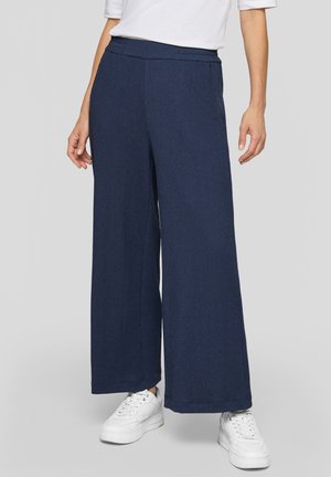 Person trägt marineblaue weit geschnittene Hose, weiße Sneakers und ein weißes kurzärmeliges Oberteil und steht vor einem schlichten hellen Hintergrund.