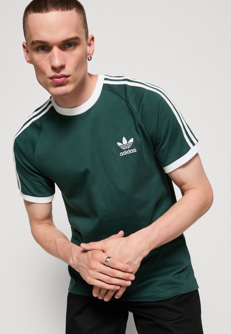 T-shirt en coton vert avec col et manches côtelés blancs. Présente le logo emblématique d'Adidas sur la poitrine et un design distinctif à trois bandes sur les épaules.