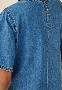 Chemise en denim à manches courtes de couleur bleu moyen. Fabriquée en denim texturé avec une couture au dos et un col. Présente des coutures traditionnelles le long des bords.