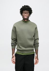 Ελιά πράσινη φόρμα με στρογγυλή λαιμόκοψη και μακριά μανίκια, κατασκευασμένη από λείο υλικό. Φορεμένη πάνω από ένα σκούρο πράσινο πουκάμισο, με ετικέτα στο μανίκι.