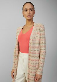 Strickjacke in Creme mit einem Wellenmuster in Pink und Grün. Kombiniert mit einem korallenfarbenen V-Ausschnitt-Top und hoch taillierten weißen Hosen.