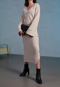 Set di cardigan e abito a coste beige con scollo a V, dettagli con bottoni laterali e spacco alto. Abbinato a stivaletti neri alla caviglia.