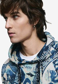 Mikina s kapucňou s modro-krémovým grafickým vzorom, mäkkou textúrou, šnúrkou a čiernymi detailmi, nosená cez biele tričko.