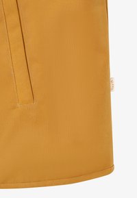 Lähikuva sinapinkeltaisesta kankaasta, jossa on pystysuora taskusauma ja pieni valkoinen brändilappu sivussa.