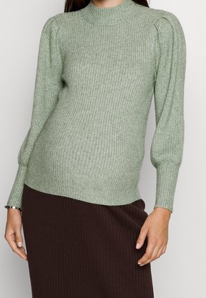 Kvinde iført en lysegrøn ribstrikket sweater med oppustede skuldre og lange ærmer, kombineret med et mørkebrunt ribstrikket nederdel.