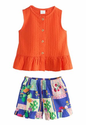 Oranžové textúrované tielko bez rukávov s gombíkmi vpredu a volánovým lemom v kombinácii s farebnými krátkymi nohavicami s kvetinovou záplatou a volánovým okrajom.