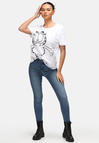 T-shirt blanc avec un motif de chat en cartoon noir, associé à un jean skinny bleu et des bottines noires à lacets. Tenue simple et décontractée.