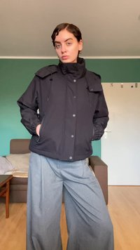 Black, lightweight jacket featuring a high collar, front buttons, and adjustable drawstrings. Paired with grey wide-leg trousers.