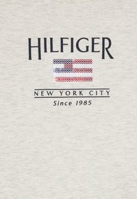 Šedé bavlněné tričko s nápisem "HILFIGER" nad texturovaným designem americké vlajky. Pod tím je nápis "NEW YORK CITY" a "Since 1985" v černém písmu.