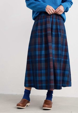 Person mit blauem Pullover, langem blau-braun kariertem Rock, blauen Socken und braunen Lederclogs, die auf einem grauen Boden steht.