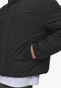 Sort polstret jakke med en lynlås lomme, en glat tekstur og et logo på brystet. Forlænget med en hvid T-shirt under.