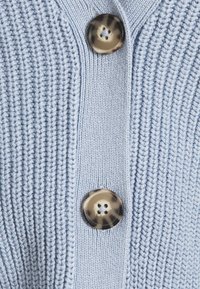 Ljusblå stickad kofta med ribbad textur, prydd med en framträdande mönstrad brun knapp.