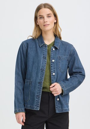 Frau trägt eine blaue Jeansjacke über einem grünen Pullover und schwarzen Hosen, steht vor einem schlichten hellen Hintergrund.