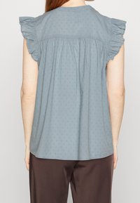 Blusa di cotone azzurro chiaro con maniche corte a volant, schiena arricciata, con piccoli e delicati motivi a pois in tutta la superficie. Texture liscia.
