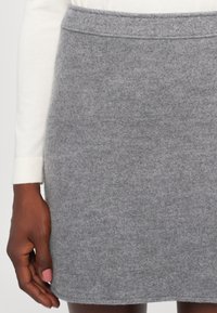 Mini-jupe en feutre gris avec une finition texturée, dotée d'une large ceinture et de détails de couture subtils sur le dessus. Main partiellement visible.