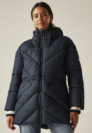 Kobieta ubrana w granatowy, pikowany zimowy płaszcz z kapturem, suwakiem z przodu i ukośnym przeszyciem, w połączeniu z niebieskimi dżinsami i złotymi kolczykami kołami.