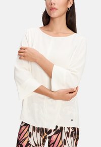Weiße Bluse mit langen Ärmeln, Bootsausschnitt und ausgestellten Ärmeln. Sie verfügt über eine subtile Nahtdetails und ein Logo-Element nahe dem Saum.