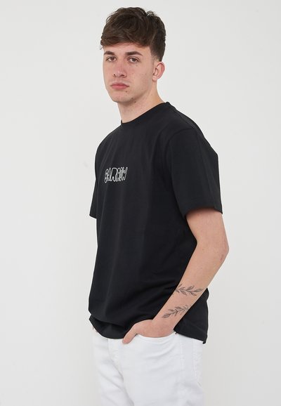 T-shirt nera in cotone con maniche corte e colletto rotondo, con un logo bianco testurizzato sul petto. Indossata con pantaloni bianchi.