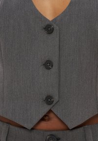 Veste grise sur mesure avec quatre boutons noirs portée sur la peau nue, dévoilant un ventre et une vue partielle d'un pantalon assorti.