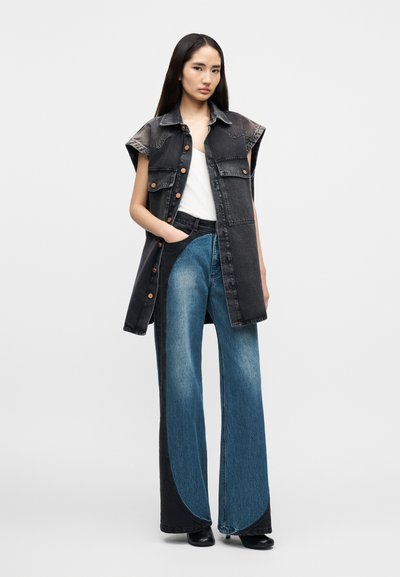 Μαύρο αμάνικο γιλέκο από τζιν με κουμπιά στο μπροστινό μέρος, φορεμένο πάνω από λευκό τοπ· συνδυασμένο με φαρδιά τζιν παντελόνια σε δυο αποχρώσεις του μπλε και μαύρα παπούτσια.