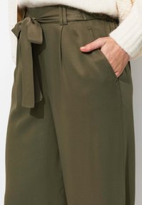 Pantalons larges vert olive avec une texture lisse, dotés d'une ceinture nouée à la taille et de poches latérales. La coupe ajustée souligne la silhouette.