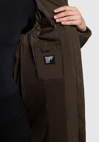 Interno di giacca marrone trapuntato con tasca per etichetta nera. Tessuto liscio e leggero; cuciture a contrasto e design semplice e funzionale.