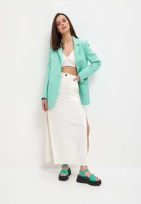 Mentolový blazer přes bílý crop top, v kombinaci s bílou sukní s bočním rozparkem a černými wedge sandály se zelenými akcenty.