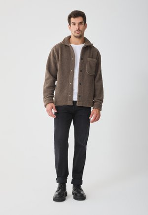Brown fleece shirt jacket with a button-down front, large chest pocket, and collar. Worn over a white shirt and paired with black jeans and shoes.