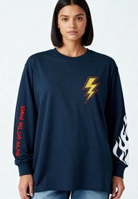 Femme portant un t-shirt à manches longues bleu marine avec un éclair jaune sur la poitrine, un texte rouge sur la manche gauche et un motif de flammes blanches sur la manche droite.