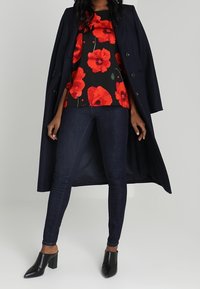 Manteau long bleu marine porté sur un haut noir imprimé de coquelicots rouges, associé à un jean skinny foncé et des chaussures noires à talons carrés.