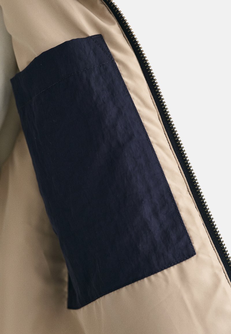 Beige Jacke mit einem dunkelblauen Stoffpatch am Ärmel. Verfügt über einen schwarzen Reißverschluss und eine glatte Textur am Körper. Das Patch hat einen leichten Glanz.