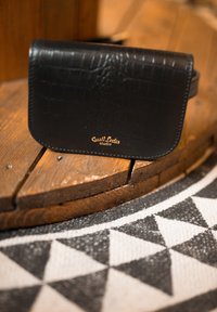 Mała czarna skórzana torebka z teksturowanym wzorem i logo "Gusti Leder studio", spoczywająca na zakrzywionej drewnianej powierzchni powyżej geometrycznego czarno-białego dywanu.