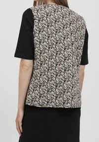 Kobieta w bezrękawnik z kwiatowym wzorem na czarnym topie z krótkim rękawem, pokazana z tyłu na jednolitym tle.