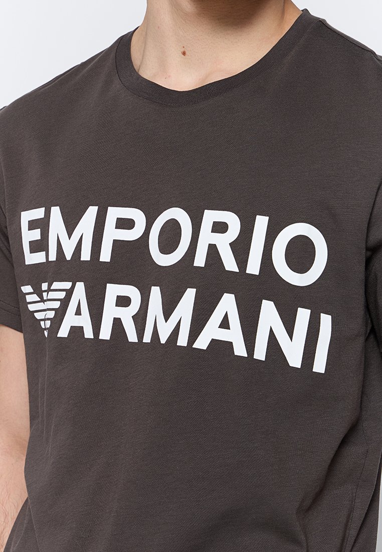 Camiseta de algodón gris oscuro con gran texto blanco "EMPORIO ARMANI" y un diseño de logo triangular en la parte delantera. Cuello redondo, mangas cortas.