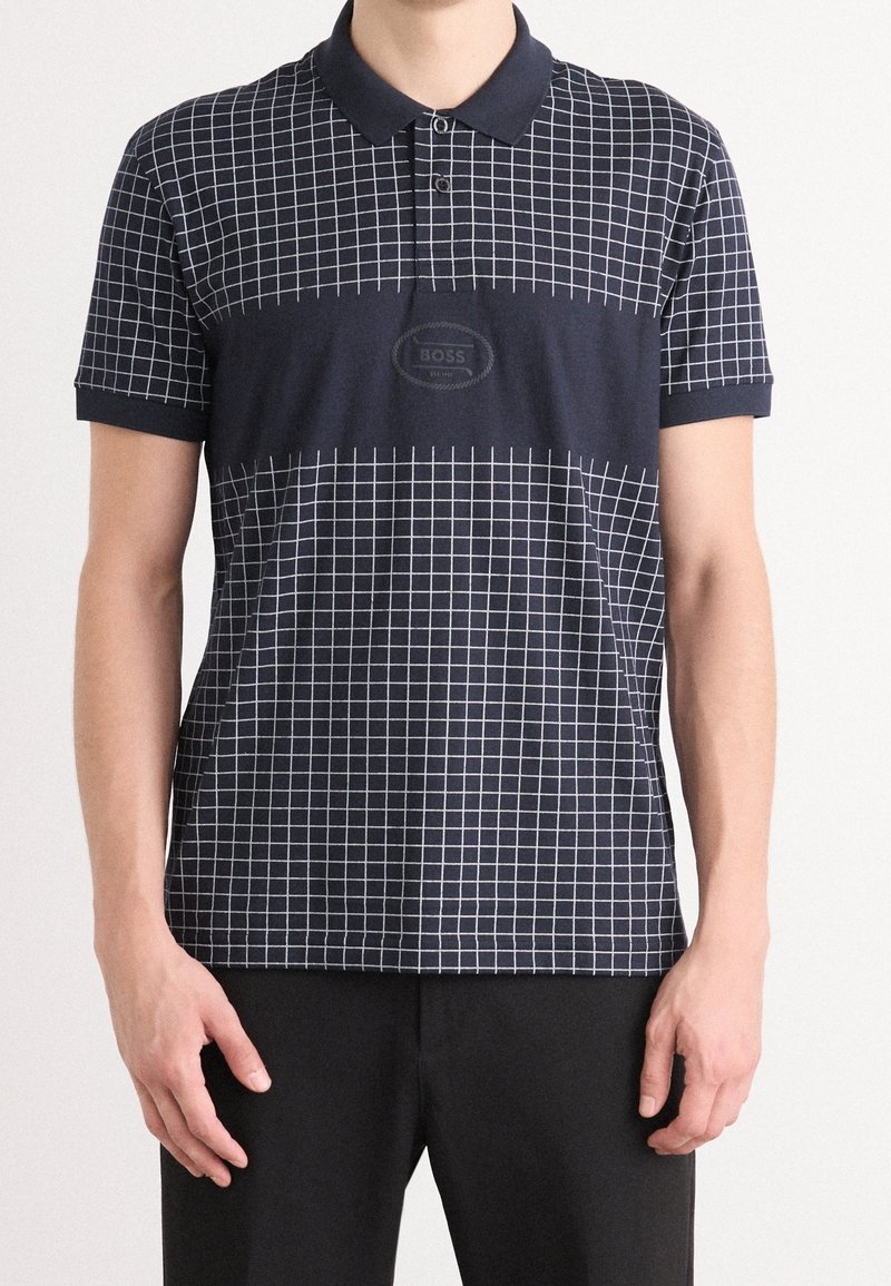Homme portant un polo bleu marine avec un motif de grille blanche et une bande centrale plus foncée avec le logo embossé "BOSS", associé à un pantalon noir.