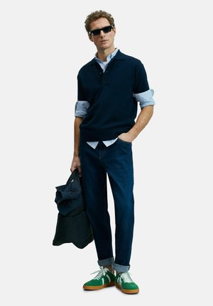Marineblauwe korte mouwen trui over een blauwgestreept shirt, donkere jeans en groene sneakers met witte veters en bruine zolen, terwijl hij een donkere jas vasthoudt.