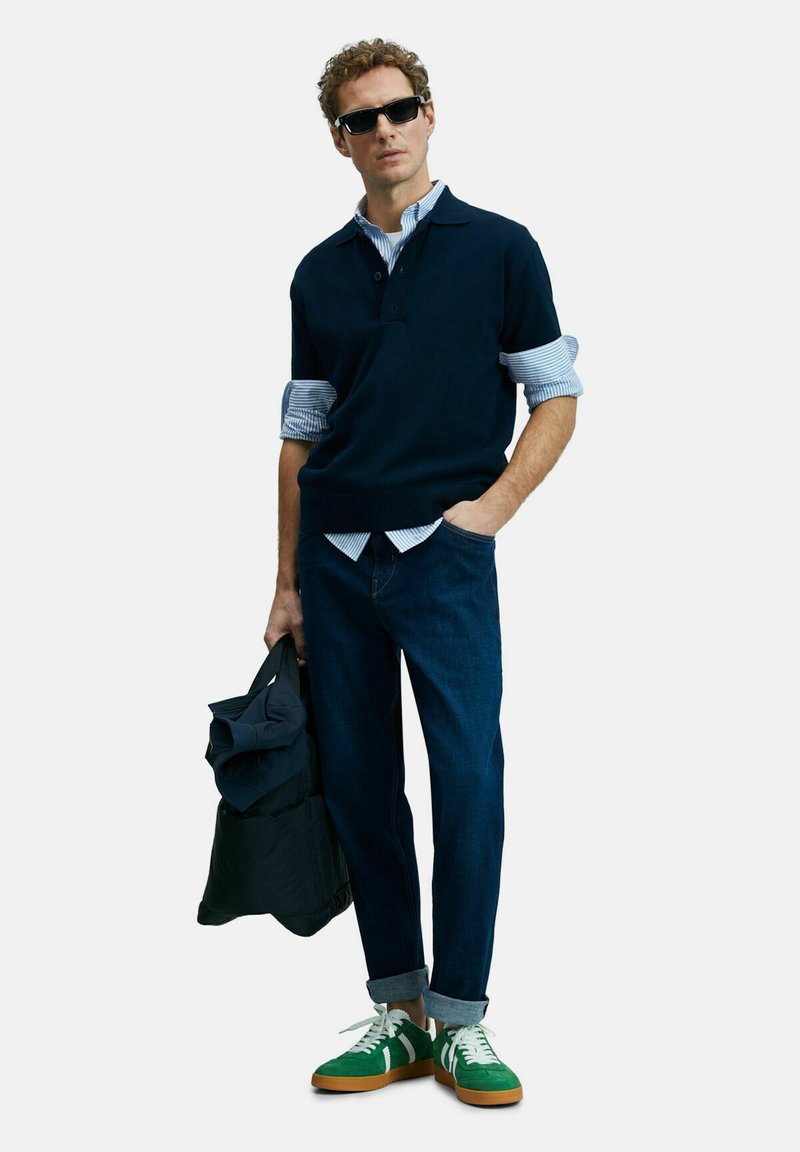 Marineblauer Kurzarmpullover über einem blau gestreiften Hemd, dunklen Jeans und grünen Sneakers mit weißen Schnürsenkeln und braunen Sohlen, während er eine dunkle Jacke hält.