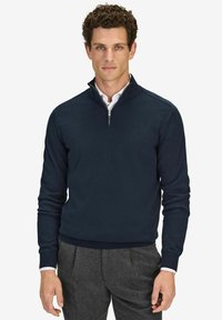 Tummansininen, vetskareilla varustettu puuvillaneulepullovaihtoehto. Varustettu kauluksella sekä resorilla varustetuilla hihoilla ja helmalla. Rikkaita valkoisen paidan päällä.